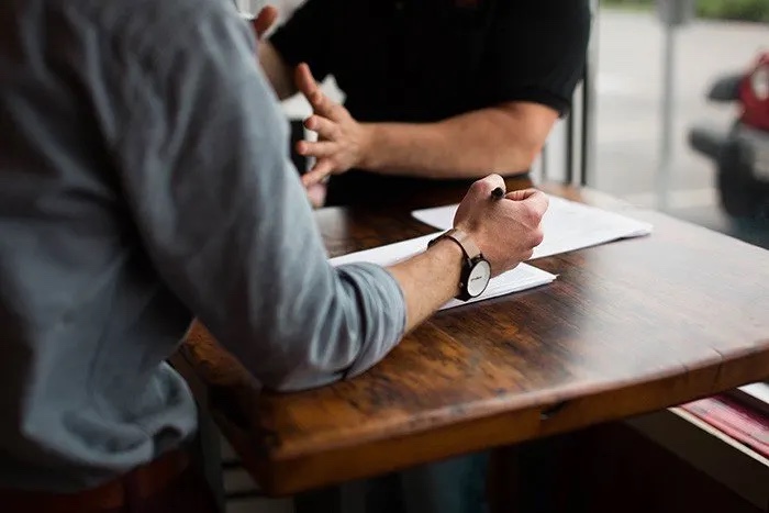 Two people are engaged in a discussion at a wooden table, with one person gesturing and the other writing on paper.