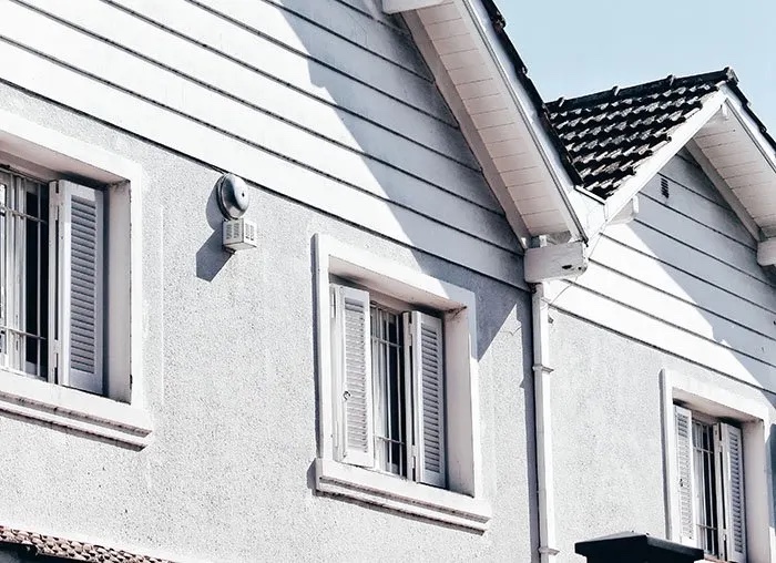 A close-up of a house exterior featuring white walls, windows with shutters, and a sloped roof under a clear sky.
